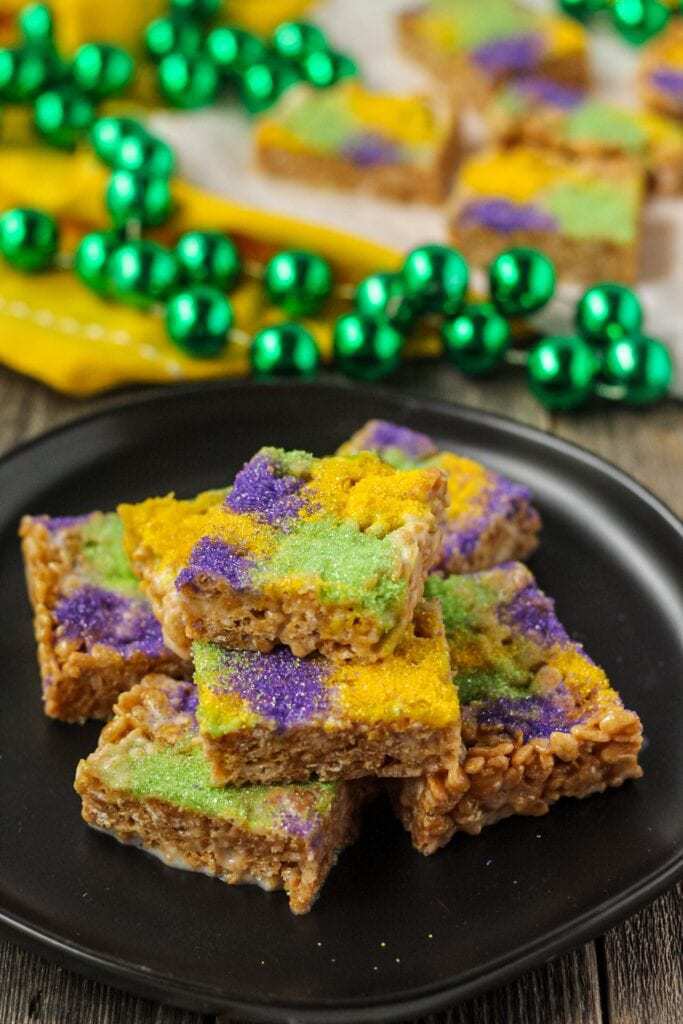 side shot of rice krispy treats made with marshmallows, butter, cinnamon and brown sugar topped with green, yellow and purple sanding sugar. treats are on a black plate and there are mardi gras beads in the background of the shot