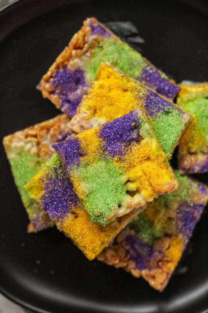 overhead shot of rice krispy treats made with marshmallows, butter, cinnamon and brown sugar topped with green, yellow and purple sanding sugar. treats are on a black plate.