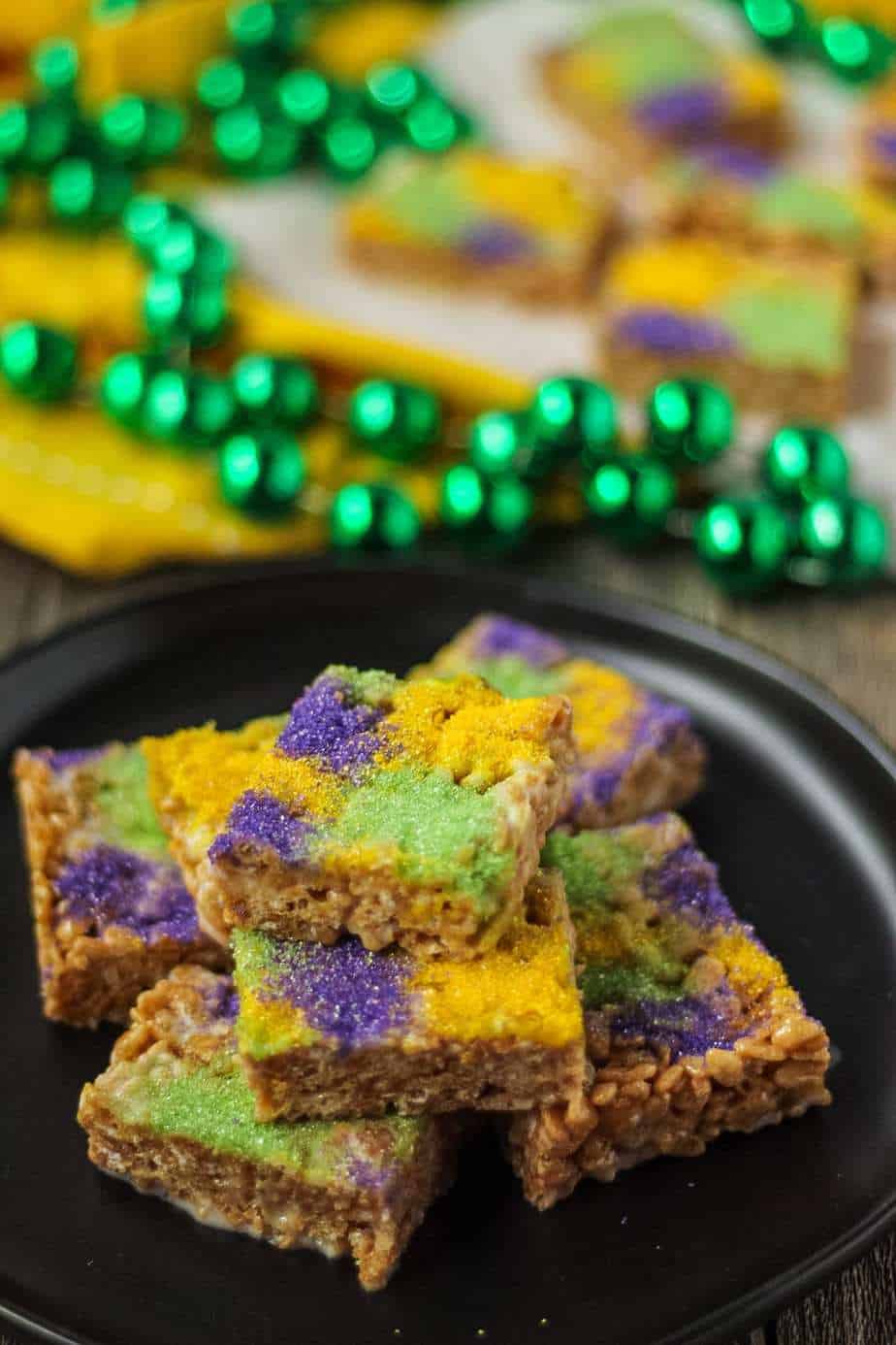 side shot of rice krispy treats made with marshmallows, butter, cinnamon and brown sugar topped with green, yellow and purple sanding sugar. treats are on a black plate and there are mardi gras beads in the background of the shot