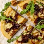overhead shot of flatbread topped with sliced pears, caramelized onions and brie cheese. Garnished with arugula and honey. Flatbread is on top of parchment on a wooden serving board.