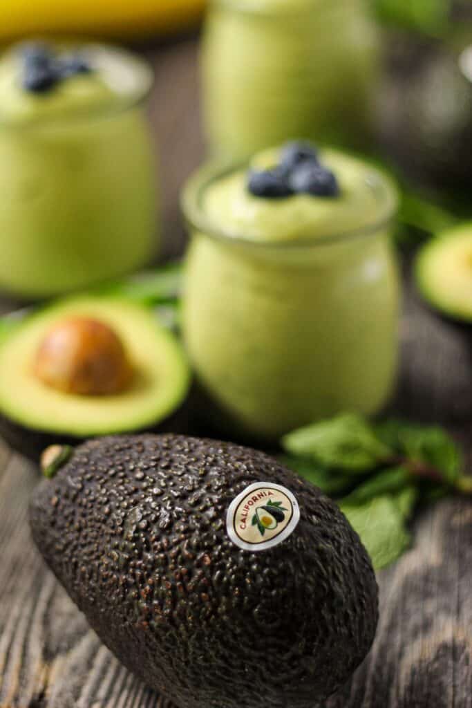 avocado smoothies in clear glasses on wood surface with a whole avocado in front of shot 