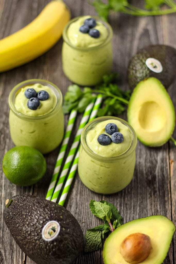 side shot of tropical avocado smoothies in clear glasses on wooden surface topped with fresh blueberries.