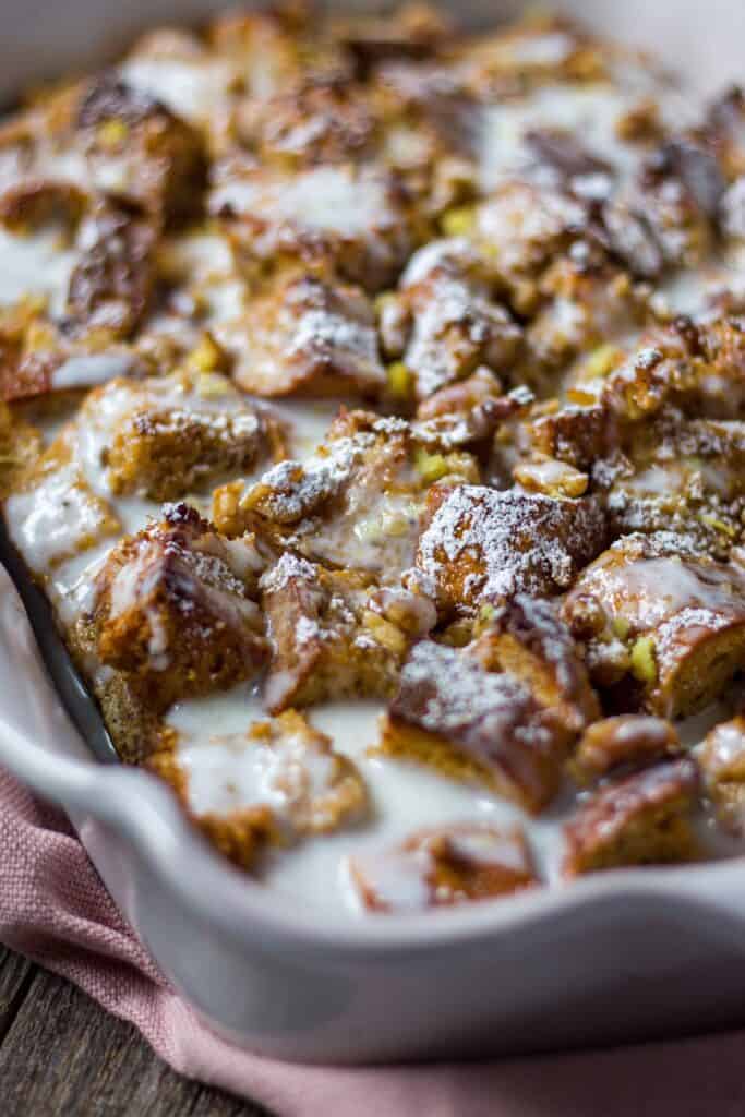 side shot of lemon bread pudding with lemon greek yogurt glaze in a terra cotta baking dish on wooden surface.