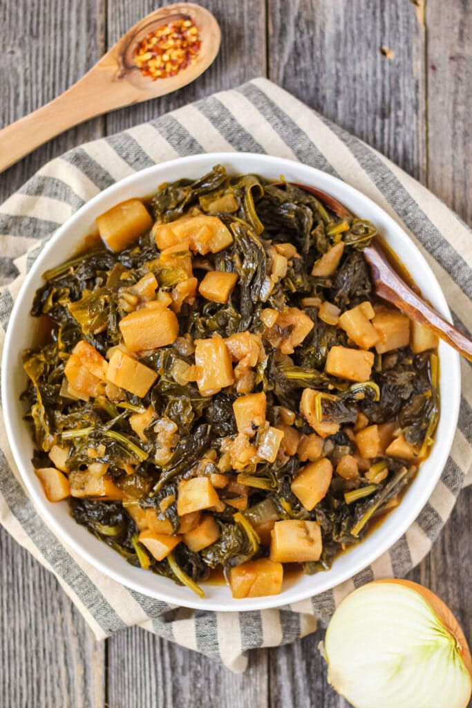 overhead shot of sweet and spicy turnip greens in white bowl on wooden surface.