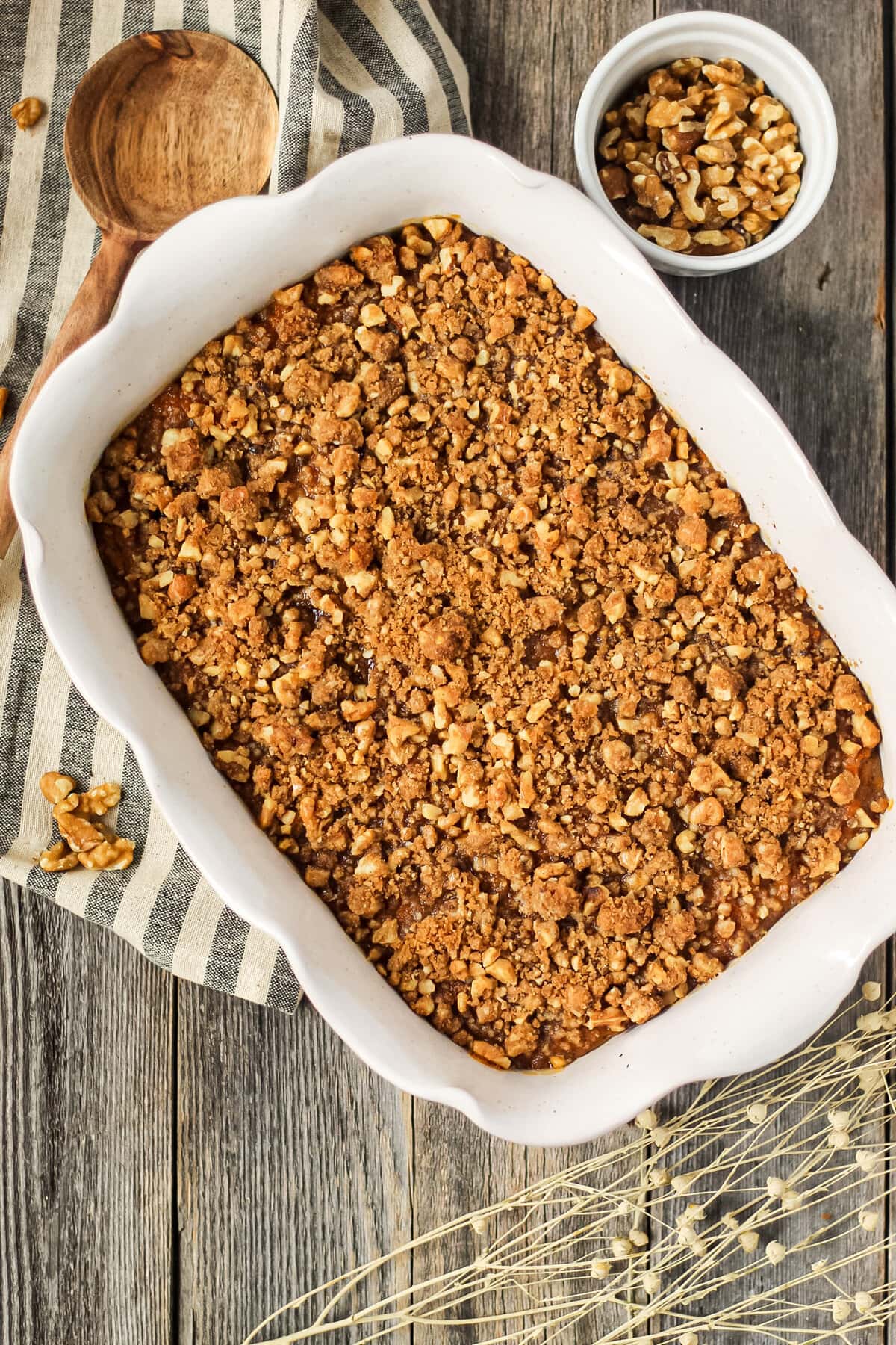 overhead shot of maple walnut sweet potato casserole in a glass baking dish on top of a wooden surface. Striped dish towel underneath dish and wooden spoon in the top left hand corner.