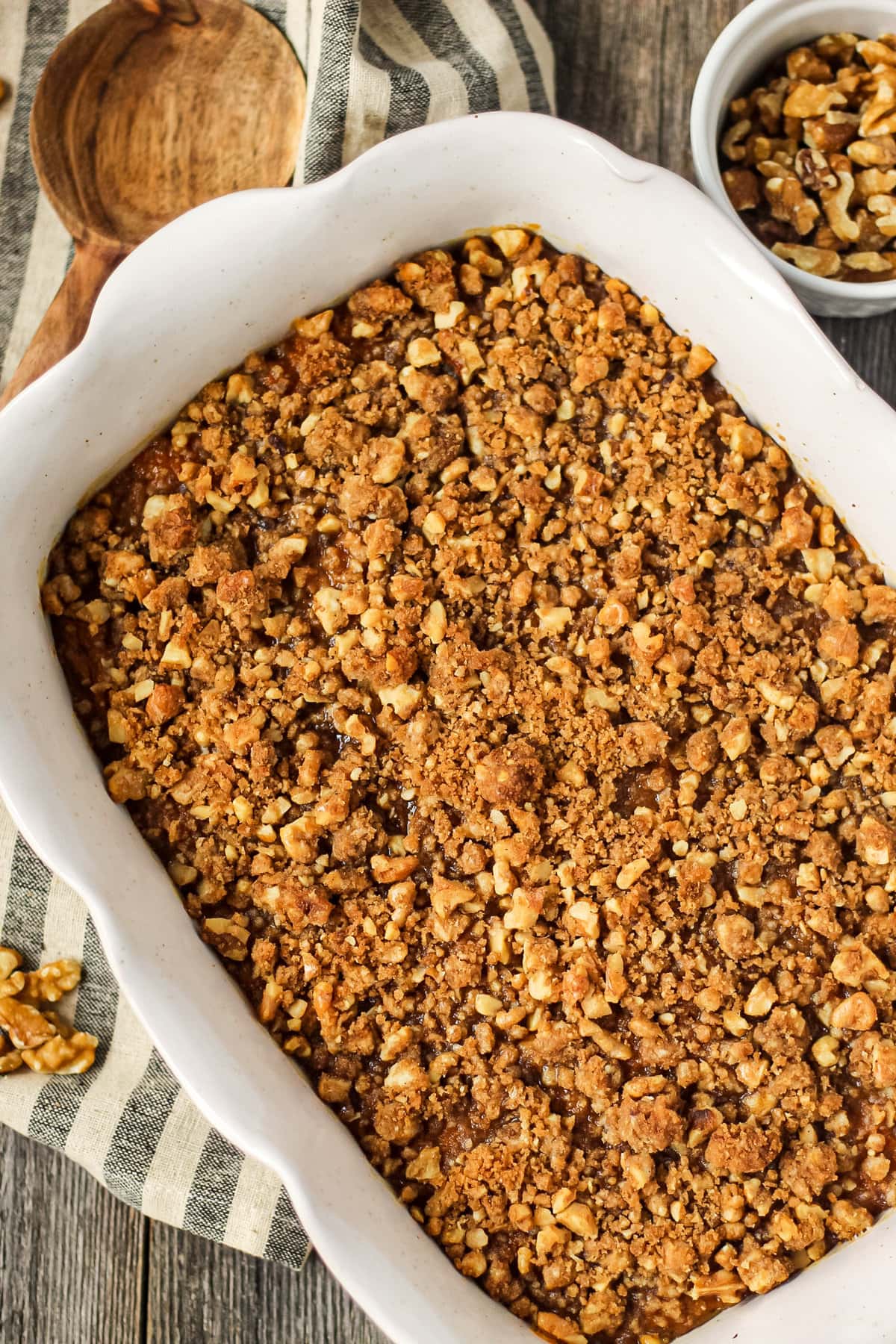overhead shot of maple walnut sweet potato casserole in a glass baking dish on top of a wooden surface. Striped dish towel underneath dish and wooden spoon in left hand corner.