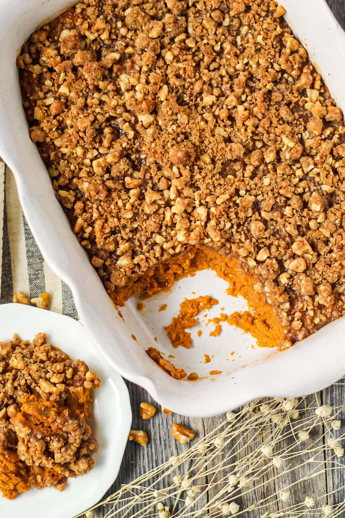 overhead shot of maple walnut sweet potato casserole in a glass baking dish on top of a wooden surface. Striped dish towel underneath dish and a small serving of casserole on a small white plate in left hand bottom corner.