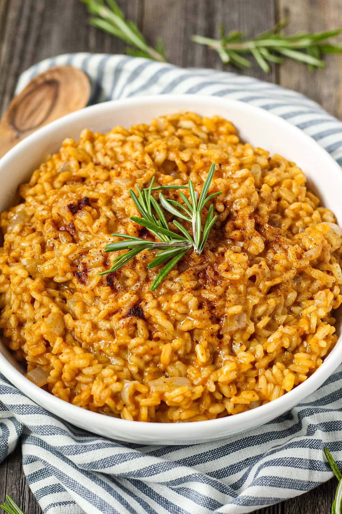 side angle shot of creamy pumpkin risotto in a white serving bowl with a striped linen underneath and a wooden serving spoon in the top left corner.