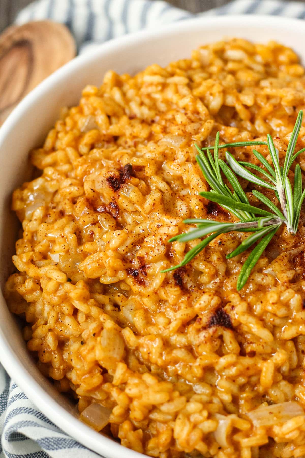 close up side angle shot of creamy pumpkin risotto in a white serving bowl with a striped linen underneath and a wooden serving spoon in the top left corner.