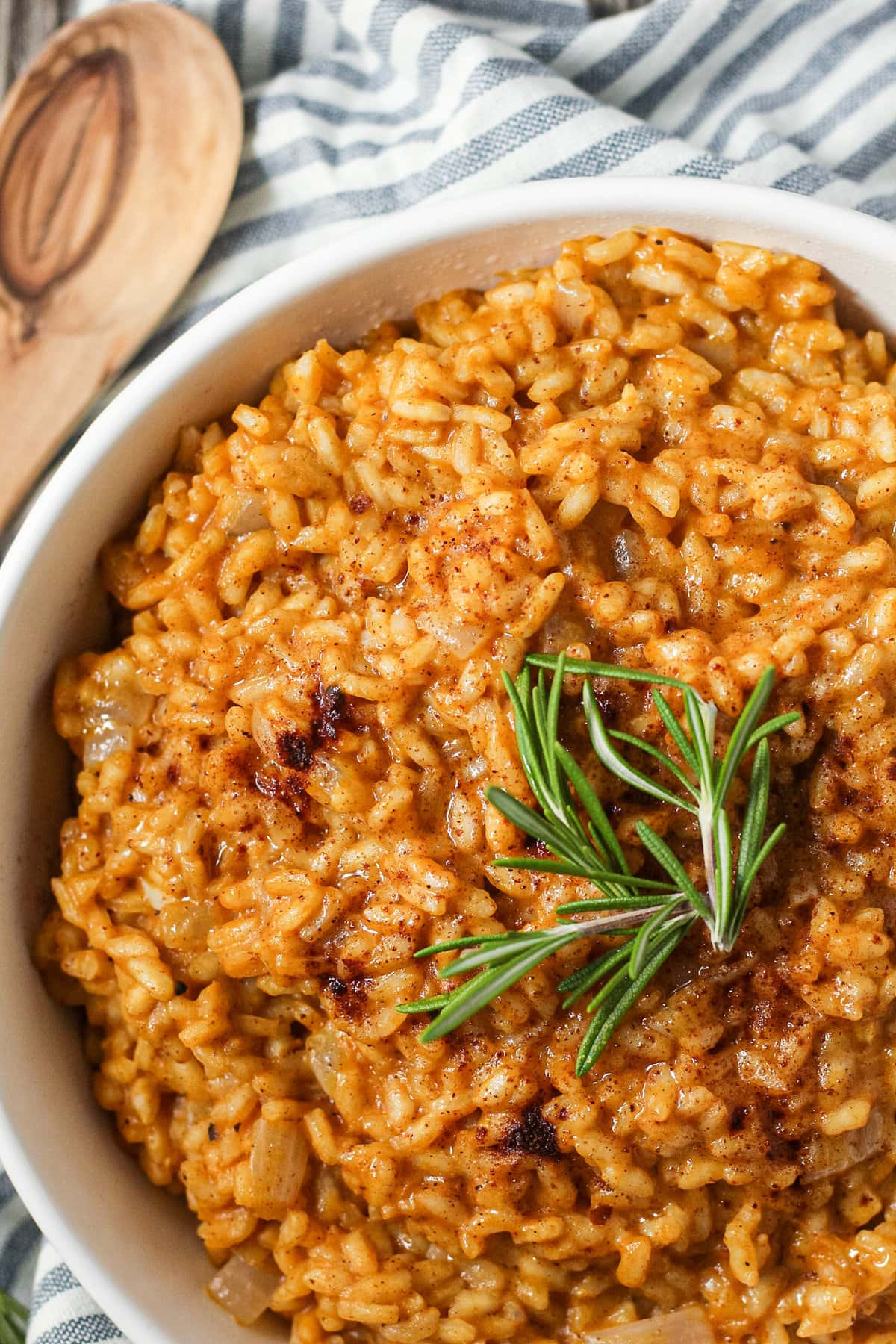 close up overhead shot of creamy pumpkin risotto in a white serving bowl with a striped linen underneath and a wooden serving spoon in the top left corner.