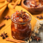 side angle shot of spiced apple jam in a glass jar with a orange linen underneath. cinnamon sticks and star anise in perimeter of shot.