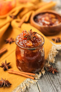 side angle shot of spiced apple jam in a glass jar with a orange linen underneath. cinnamon sticks and star anise in perimeter of shot.