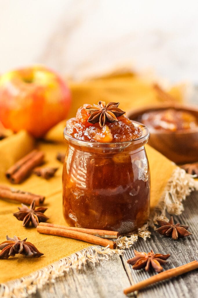 side angle shot of spiced apple jam in a glass jar with a orange linen underneath. cinnamon sticks and star anise in perimeter of shot.
