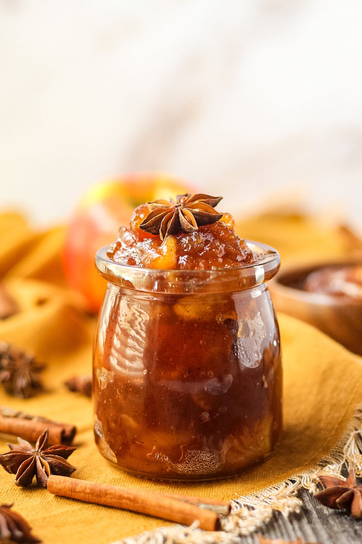 close up side angle shot of spiced apple jam in a yogurt jar with orange linen underneath and cinnamon sticks in the perimeter of the shot.