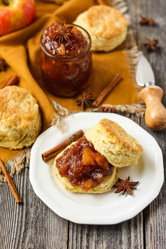 overhead shot of buttermilk biscuit sliced on a plate with spiced apple jam spread on biscuit. Plate is on a wooden surface with an orange linen underneath.