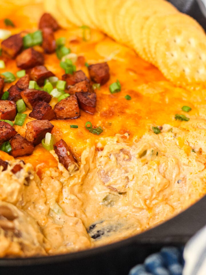 close up side angle shotof cheesy cajun chicken and sausage dip in cast iron skillet with blue towel underneath. serving of dip has been scooped out of skillet.