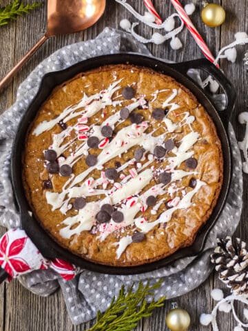 overhead shot of chocolate chunk peppermint skillet blondie in cast iron skillet on a wooden surface with a gray linen underneath skillet. copper spoon in top left corner.
