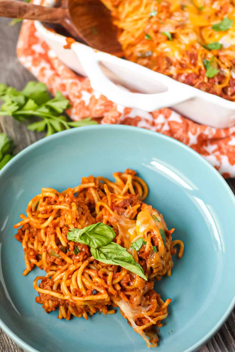 side angle shot of a serving of easy southern baked spaghetti on a blue plate on wooden surface with orange linen underneath. wooden spoon in top left corner.