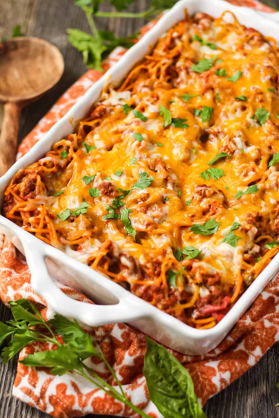 side angle shot of easy southern baked spaghetti in white baking dish on wooden surface with orange linen underneath. wooden spoon in top left corner.