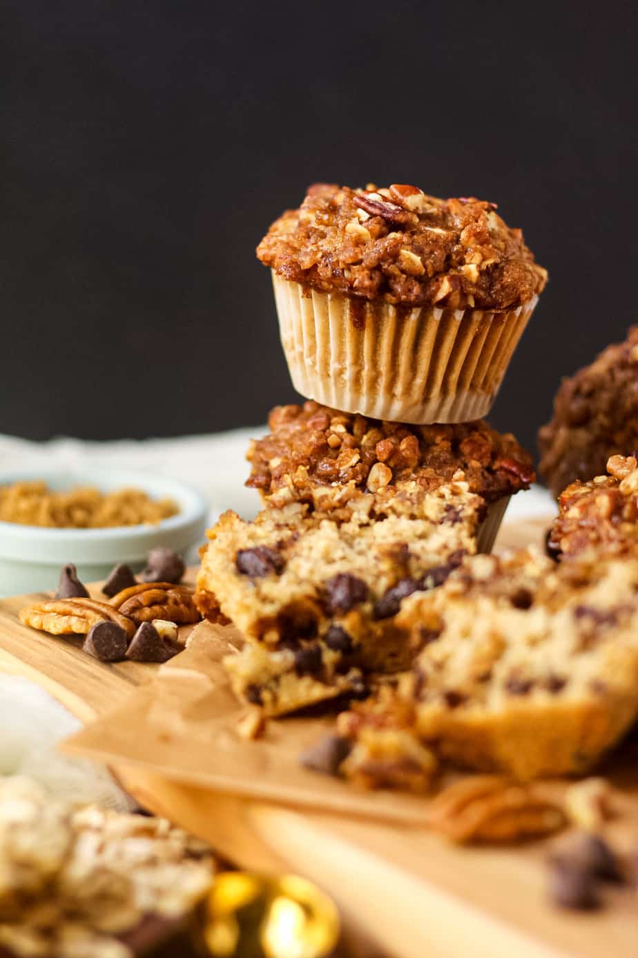 side angle shot of chocolate chip oat muffins with pecan streusel topping on a wooden cutting board sitting on a baking pan with oats and pecans in perimeter of shot.
