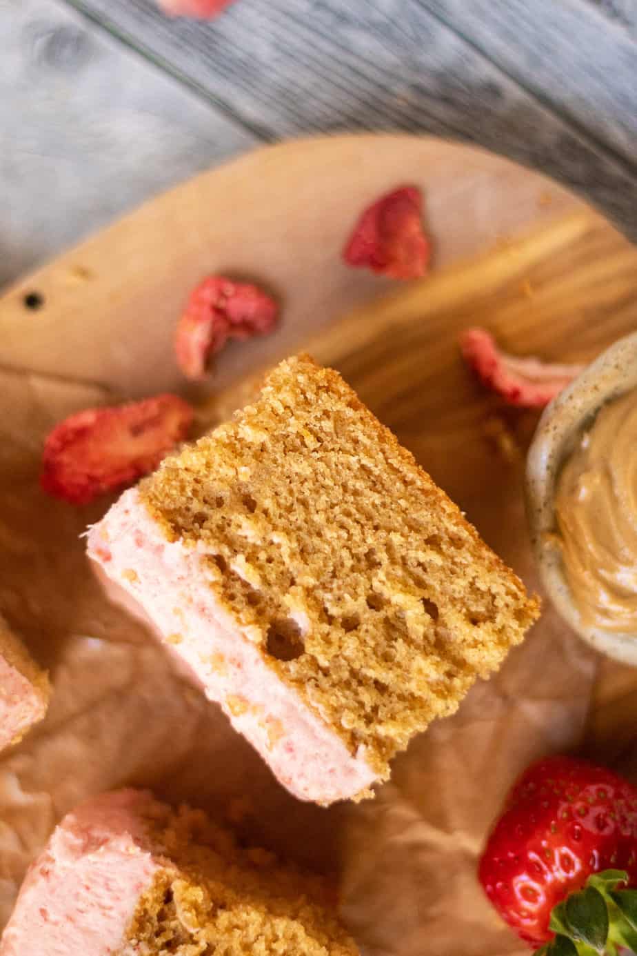 close up overhead shot of sliced peanut butter cake with strawberry frosting on a wooden cutting board. sliced strawberries scattered in perimeter of shot.