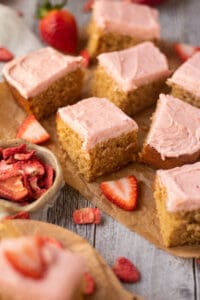 side angle shot of sliced peanut butter cake with strawberry frosting on a wooden cutting board. sliced strawberries scattered in perimeter of shot.