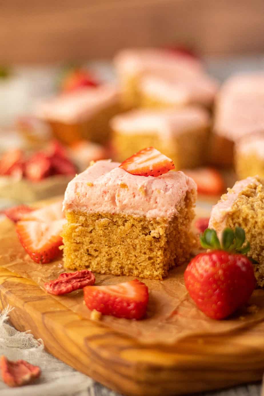side angle shot of sliced peanut butter cake with strawberry frosting on a wooden cutting board. sliced strawberries scattered in perimeter of shot.