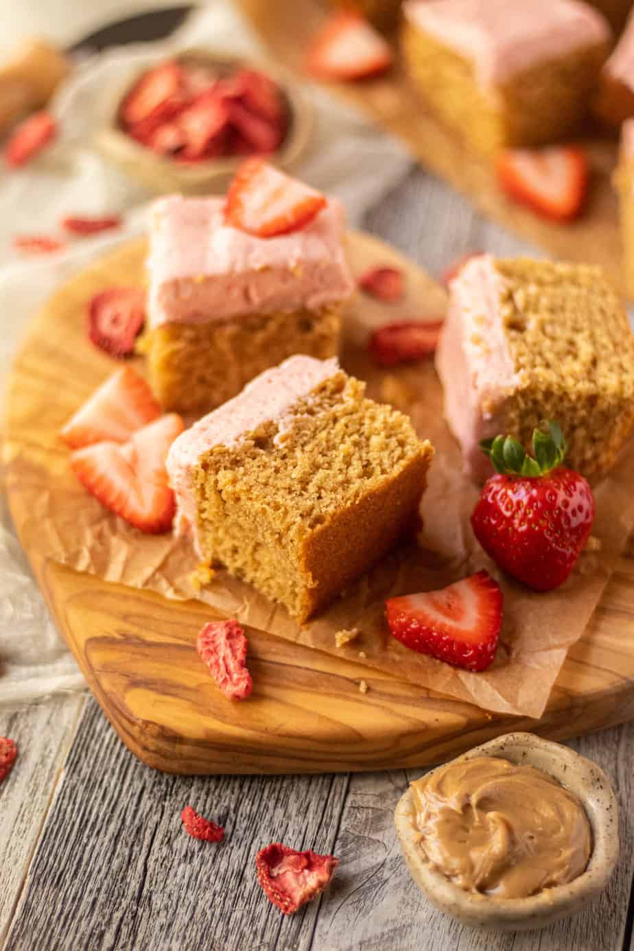 side angle shot of sliced peanut butter cake with strawberry frosting on a wooden cutting board. sliced strawberries scattered in perimeter of shot.