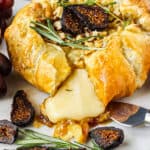 side angle close up shot of puff pastry baked brie with fig jam on white cake plate on wooden surface. grapes and bread slices around baked brie.