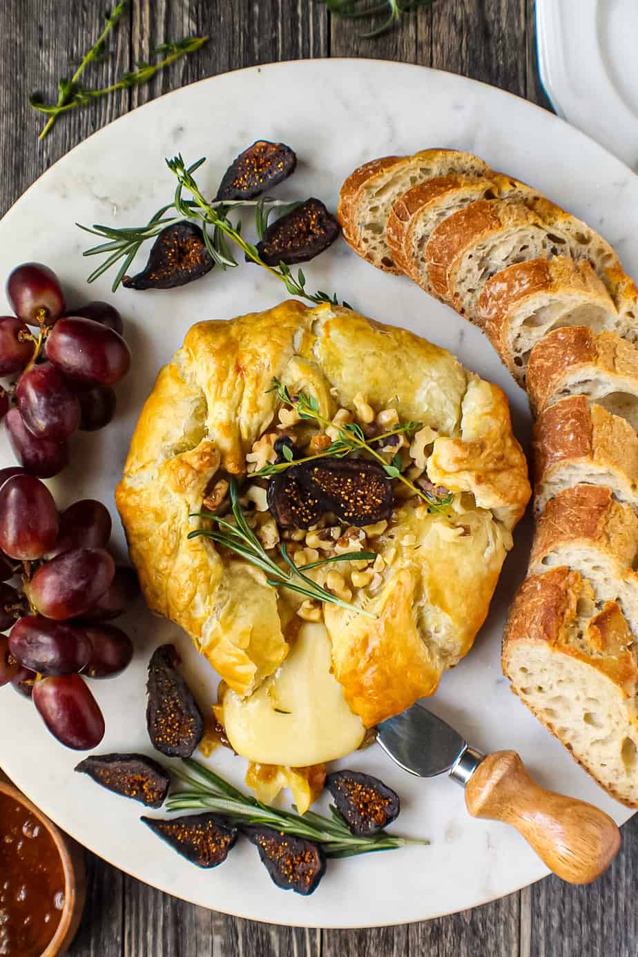 overhead shot of puff pastry baked brie with fig jam on white cake plate on wooden surface. grapes and bread slices around baked brie.
