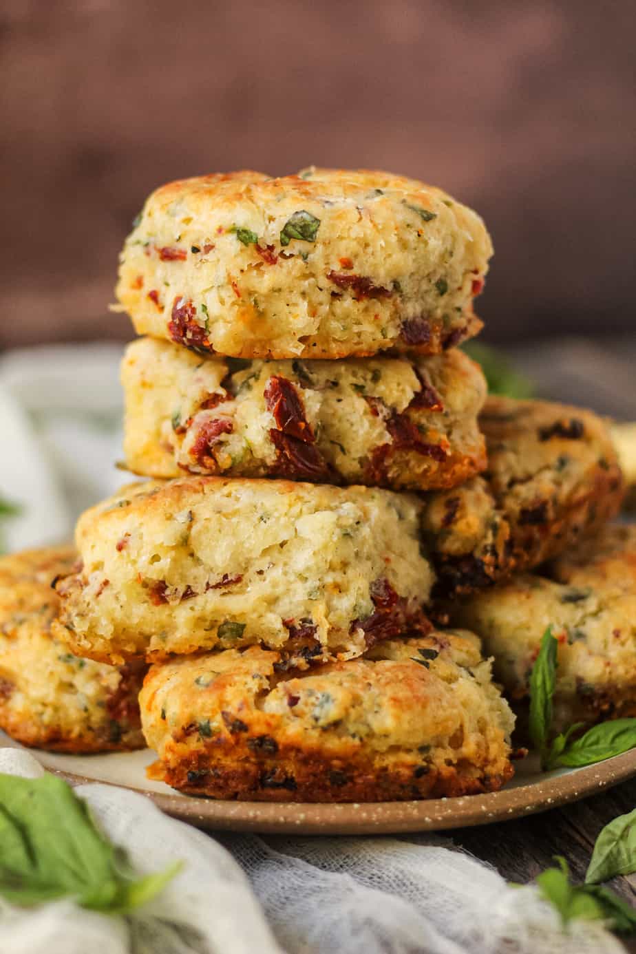 side angle shot of a stack of sun-dried tomato, cheddar, and herb biscuits on a gray plate on a wooden surface. fresh basil in perimeter of shot.