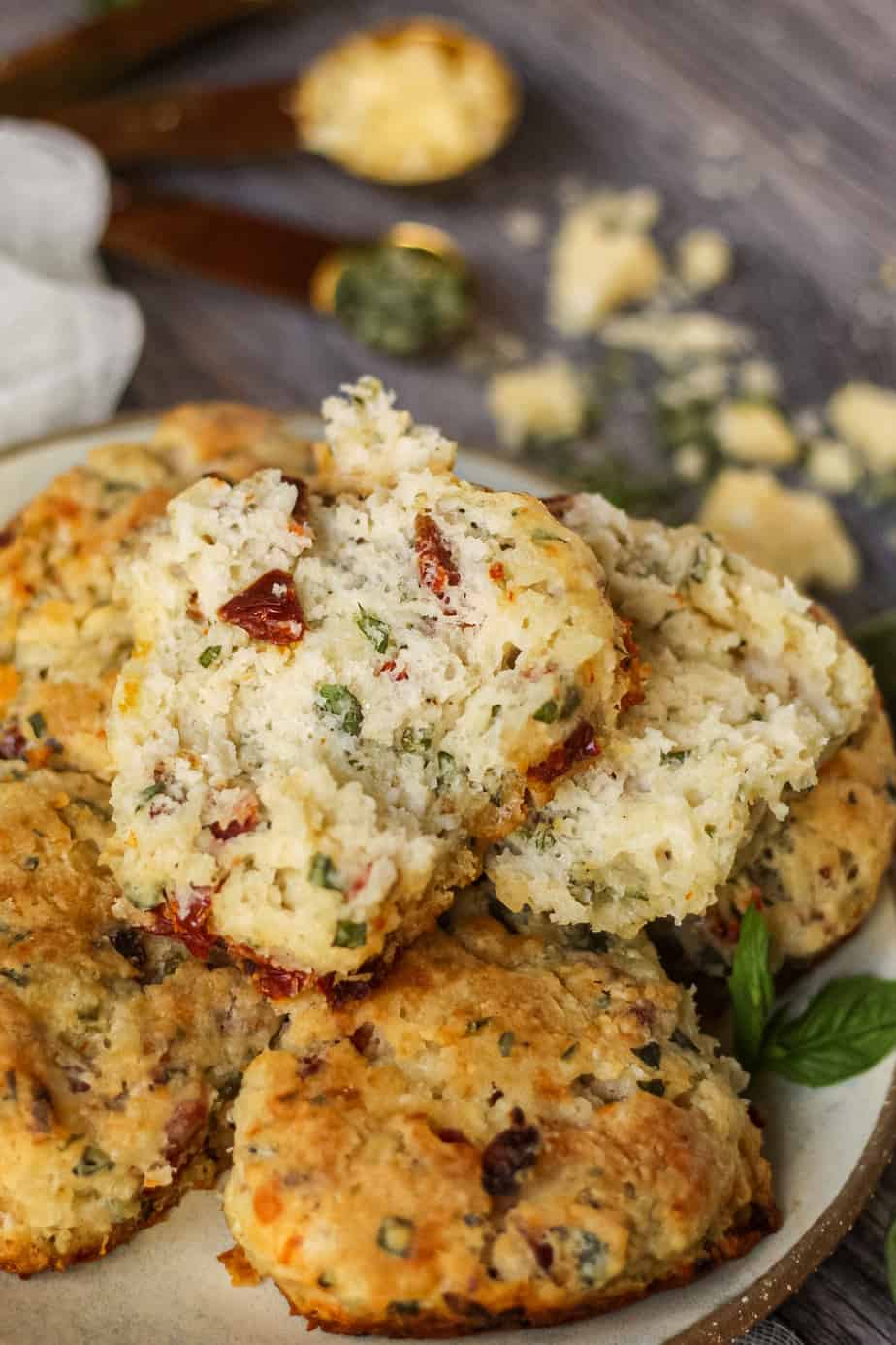 45 degree angle shot of a stack of sun-dried tomato, cheddar, and herb biscuits on a gray plate on a wooden surface. fresh basil in perimeter of shot. one biscuit broken open on top to show texture.