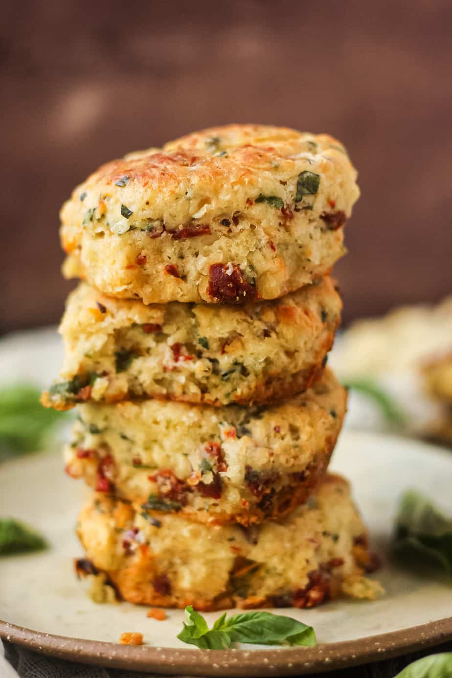 side angle shot of a stack of four sun-dried tomato, cheddar, and herb biscuits on a gray plate on a wooden surface. fresh basil in perimeter of shot.