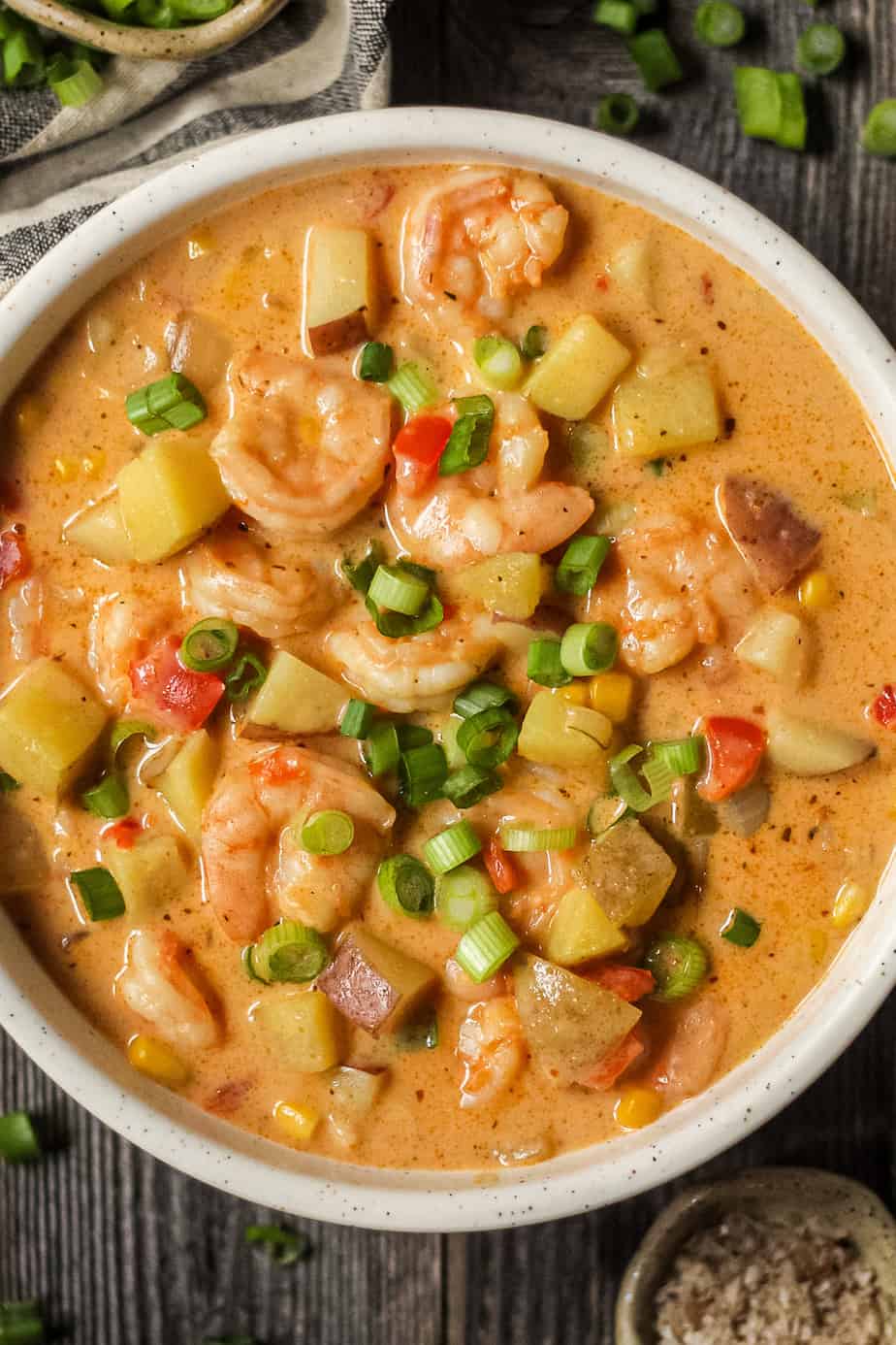 overhead shot of creamy cajun shrimp and corn chowder in a white bowl on a wooden surface. Bowl of salt and chopped green onions in top left and bottom right of shot.