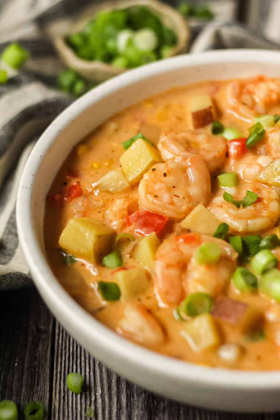 side angle shot of creamy cajun shrimp and corn chowder in a white bowl on a wooden surface. Bowl of salt and chopped green onions in top left and bottom right of shot.