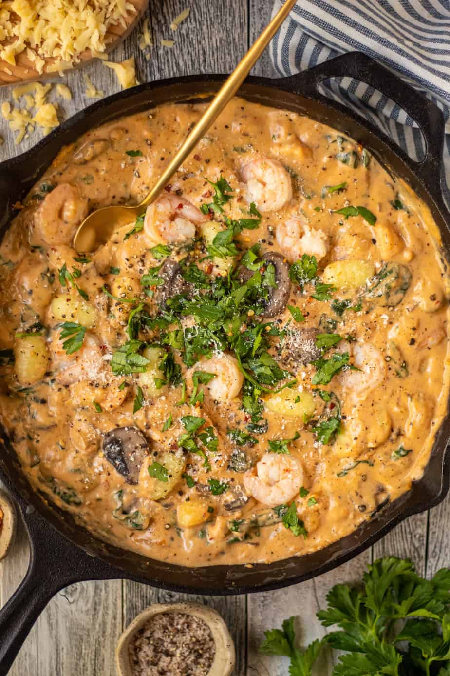overhead shot of Creamy Cajun Shrimp and Gnocchi Skillet in a cast iron skillet on top of white washed wooden surface. striped linen in perimeter.