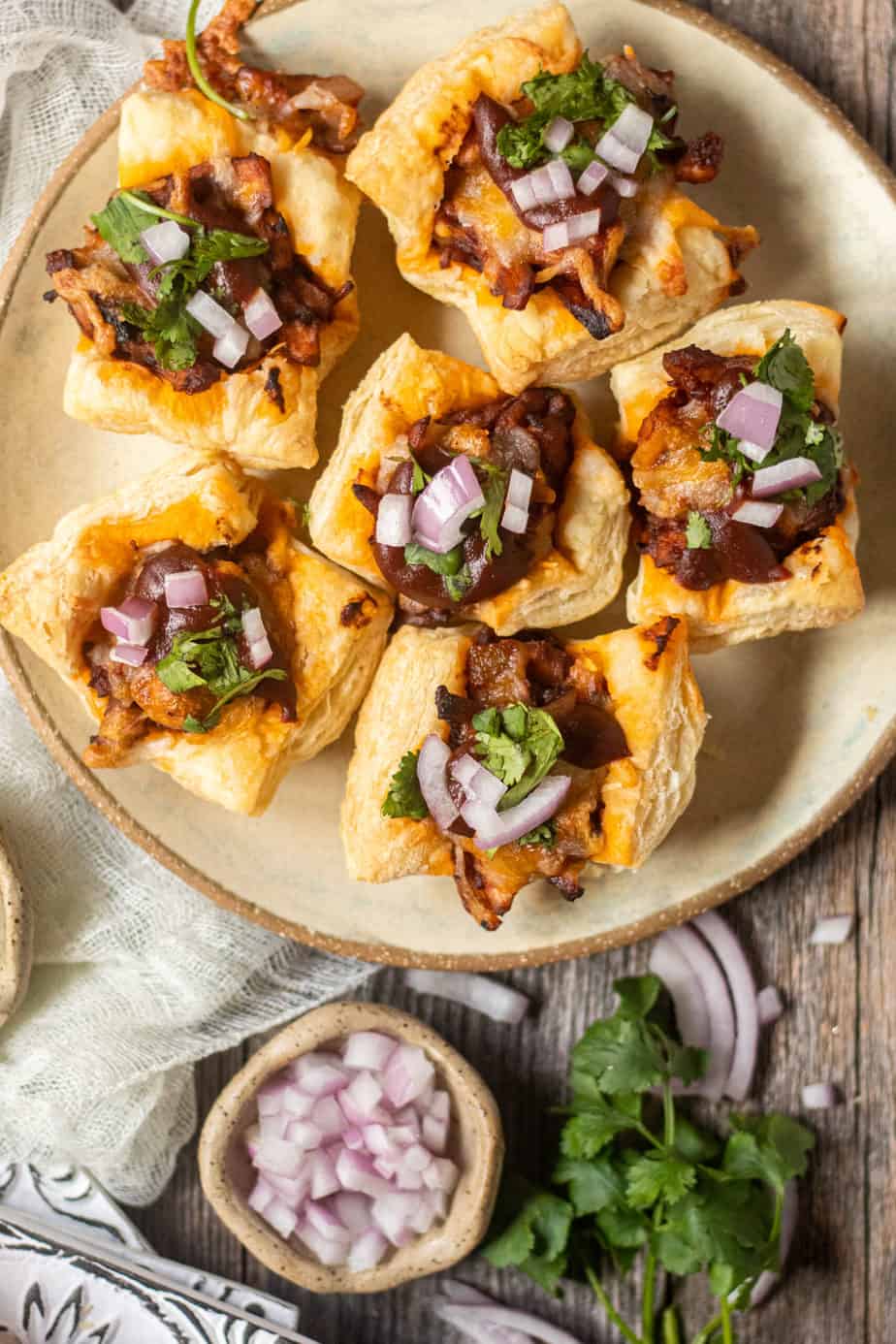 overhead shot of Sweet & spicy bbq chicken puff pastry cups on a gray plate on wooden surface. chopped red onion and cilantro in perimeter of shot
