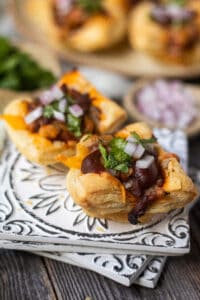 side angle shot of Sweet & spicy bbq chicken puff pastry cups on a gray plate on wooden surface. chopped red onion and cilantro in perimeter of shot
