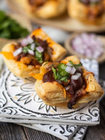 side angle shot of Sweet & spicy bbq chicken puff pastry cups on a gray plate on wooden surface. chopped red onion and cilantro in perimeter of shot
