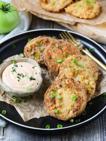 fried green tomatoes on a black plate served with creamy dipping sauce in a cup.