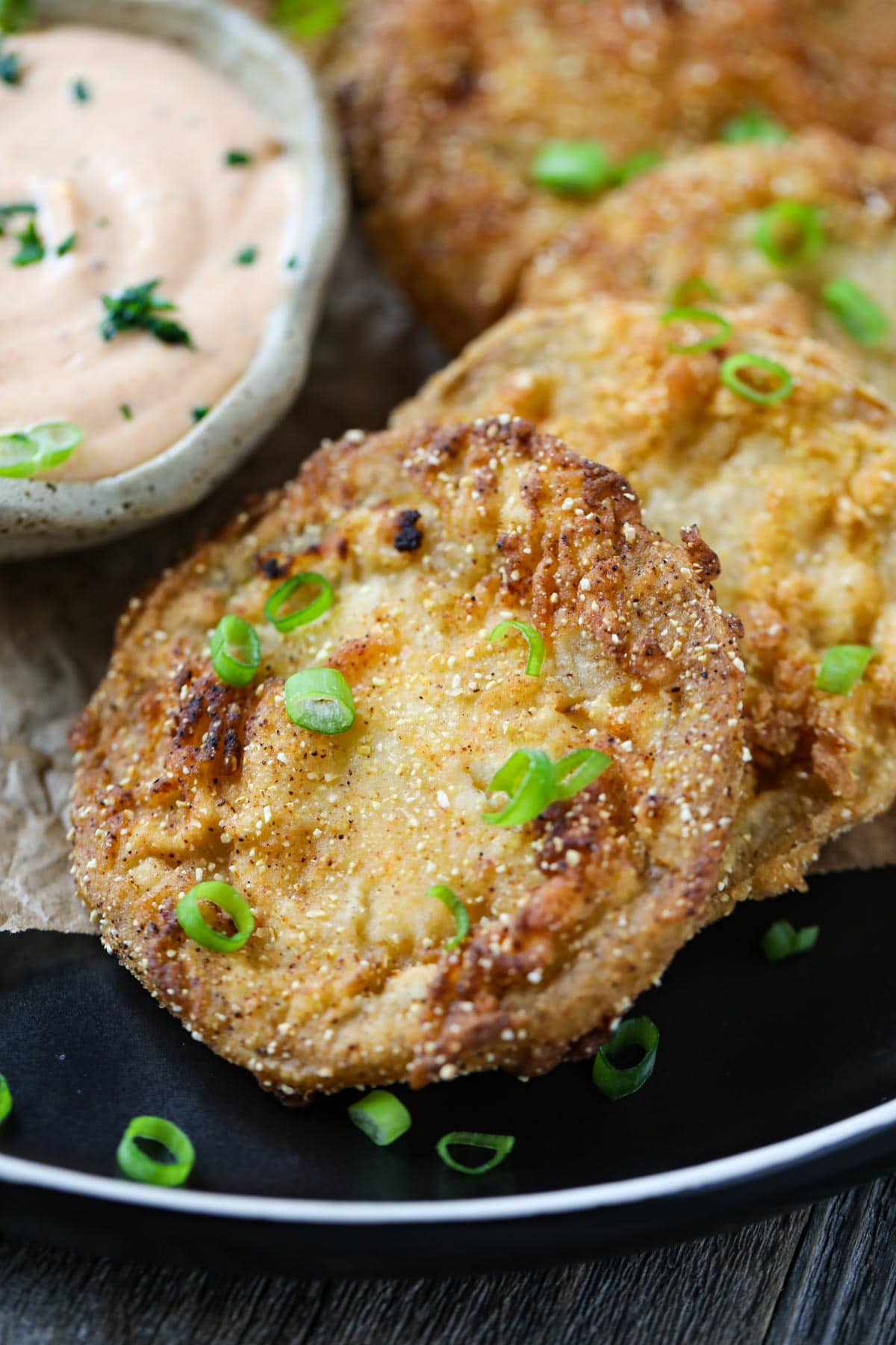 fried green tomatoes on a black plate served with creamy dipping sauce in a cup.