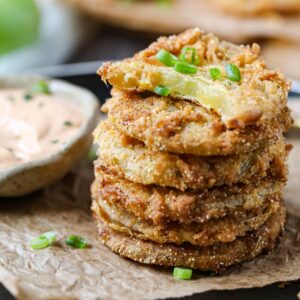 fried green tomatoes stacked on a black plate served with creamy dipping sauce in a cup.