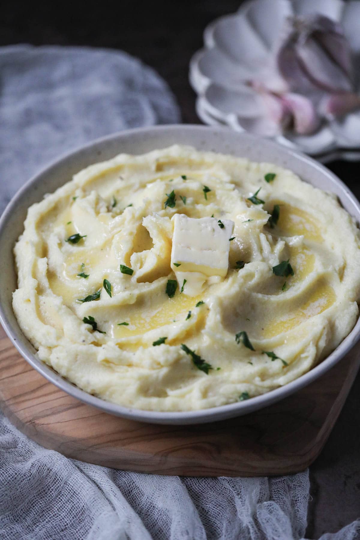 creamy garlic mashed potatoes in a white bowl with plate of garlic in the top right corner.