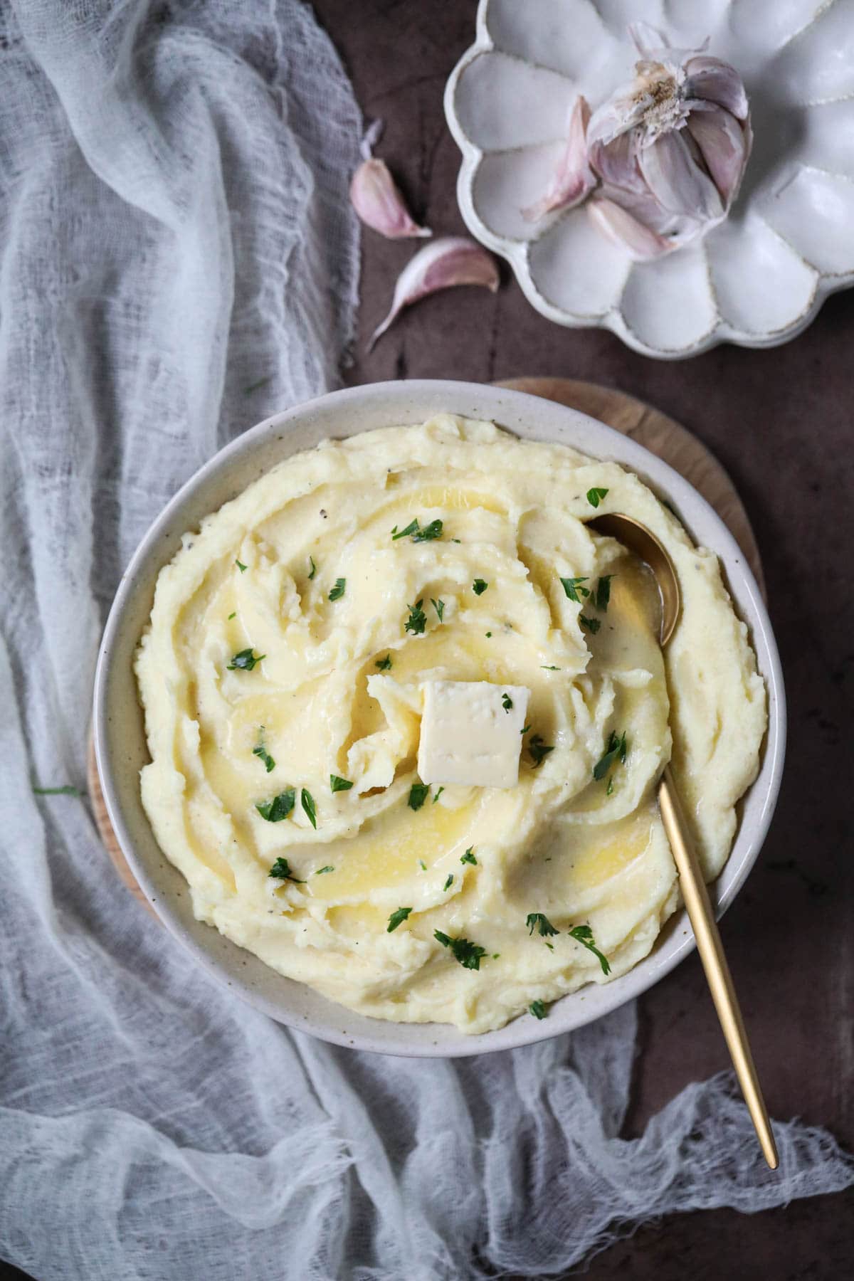 creamy garlic mashed potatoes in a white bowl with plate of garlic in the top right corner.