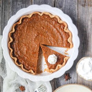 southern sweet potato pie sliced in a white pie dish and a single slice on a plate with whipped cream on a wooden surface.