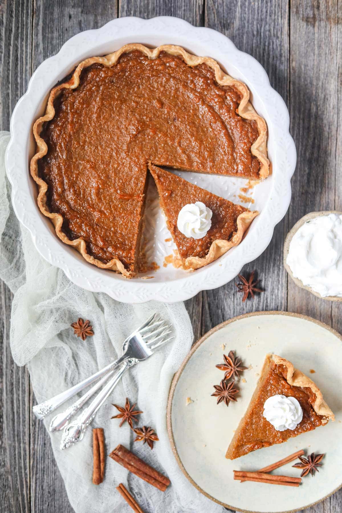 southern sweet potato pie sliced in a white pie dish and a single slice on a plate with whipped cream on a wooden surface.