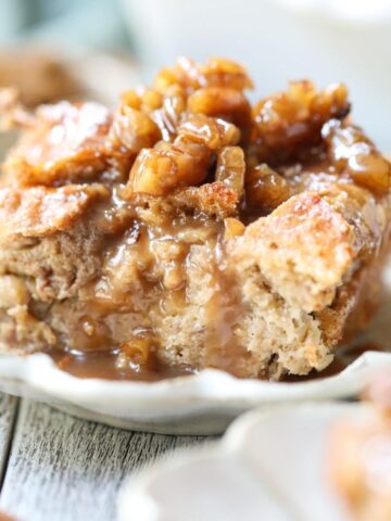 A slice of bread pudding with maple praline sauce on a white plate with a pan of bread pudding in the background.
