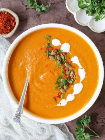 creamy sweet potato soup in a white bowl garnished with coconut cream, red pepper, and parsley. Bowl of parsley and pepper flakes in the perimeter of the shot.