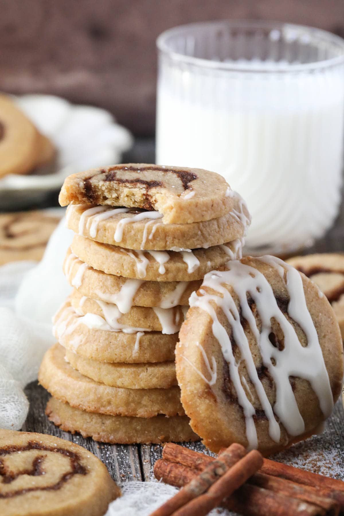 cinnamon roll cookies stacked on a wooden surface with cinnamon sticks and a glass of milk in the corner.