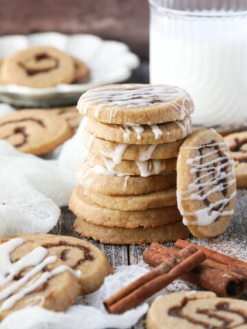 cinnamon roll cookies stacked on a wooden surface with cinnamon sticks and a glass of milk in the corner.